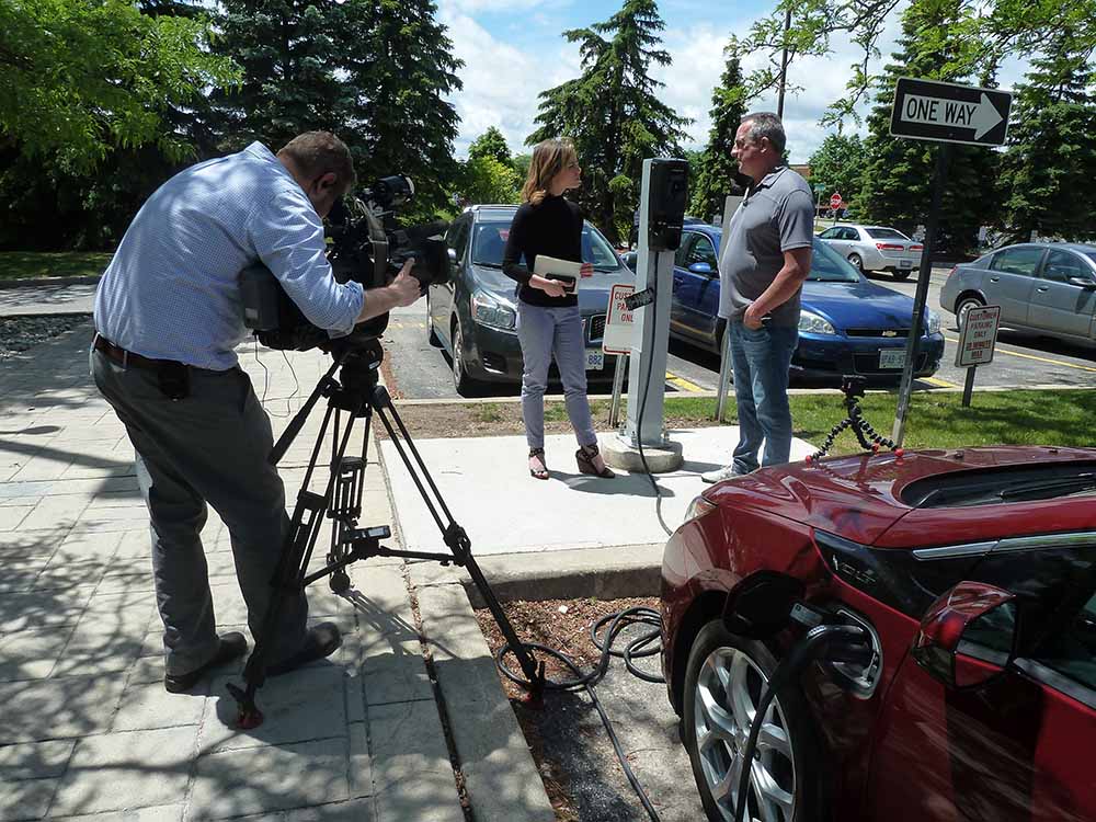 Camera man stands with camera and records two people speaking in a parking lot next to an electric vehicle charger and car.